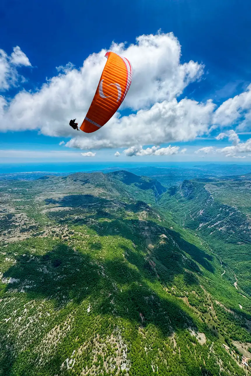 Vol parapente biplace, voile orange et pilote au-dessus des Préalpes d'Azur verdoyantes et de la Méditerranée — Ailéments Côte d'Azur